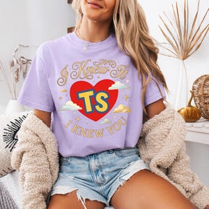 May include: A light purple short-sleeve t-shirt featuring a graphic of a red heart with the yellow letters TS inside. The text I KNEW IT is arched above the heart and I KNEW YOU is below, surrounded by small gold stars and white clouds.