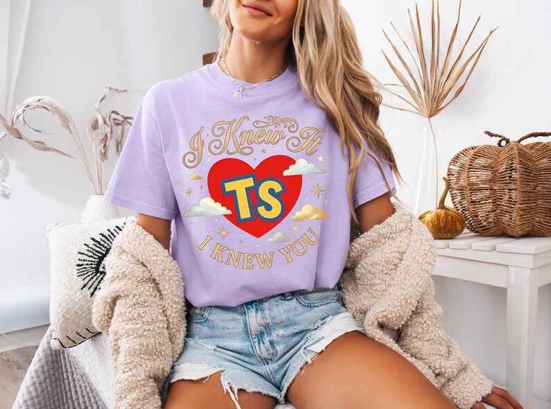 May include: A light purple short-sleeve t-shirt featuring a graphic of a red heart with the yellow letters TS inside. The text I KNEW IT is arched above the heart and I KNEW YOU is below, surrounded by small gold stars and white clouds.