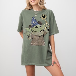 May include: An olive green short-sleeve t-shirt featuring a graphic of a green creature wearing a blue wizard hat with a white crescent moon. The creature holds a wand with a Mickey Mouse-shaped star at the tip, surrounded by small black hearts.