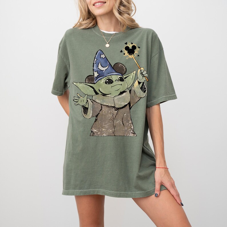 May include: An olive green short-sleeve t-shirt featuring a graphic of a green creature wearing a blue wizard hat with a white crescent moon. The creature holds a wand with a Mickey Mouse-shaped star at the tip, surrounded by small black hearts.