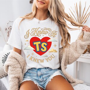 May include: A white short-sleeve t-shirt featuring a graphic design with a large red heart in the centre. Inside the heart are the yellow letters TS with a blue outline. The text I KNEW IT I KNEW YOU is printed in gold cursive font.