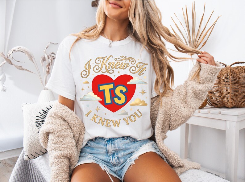 May include: A white short-sleeve t-shirt featuring a graphic design with a large red heart in the centre. Inside the heart are the yellow letters TS with a blue outline. The text I KNEW IT I KNEW YOU is printed in gold cursive font.