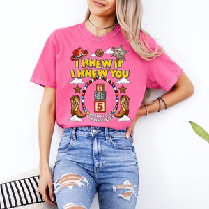 May include: A bright pink short-sleeve t-shirt featuring a graphic print with the text I KNEW IT I KNEW YOU, a cowboy hat, a sheriff badge, two cowboy boots, and a circular frame of colorful beads surrounding three stacked alphabet blocks.