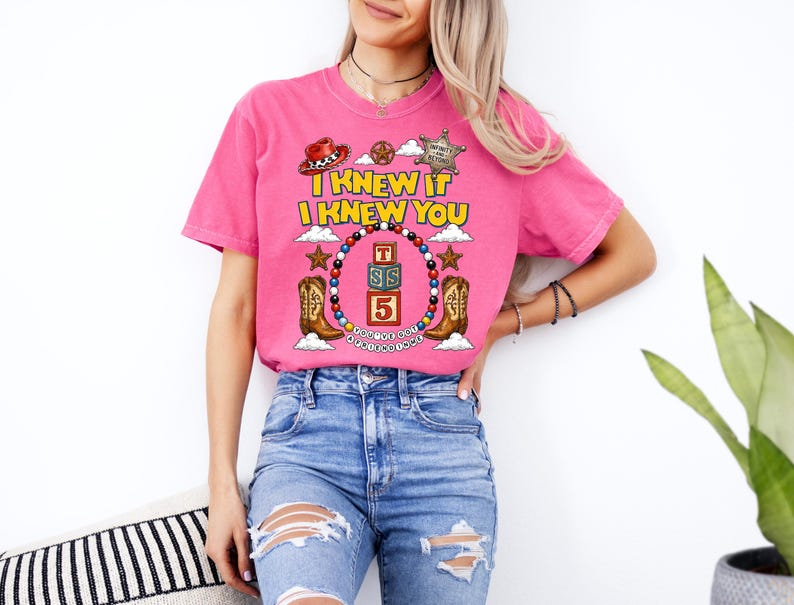 May include: A bright pink short-sleeve t-shirt featuring a graphic print with the text I KNEW IT I KNEW YOU, a cowboy hat, a sheriff badge, two cowboy boots, and a circular frame of colorful beads surrounding three stacked alphabet blocks.