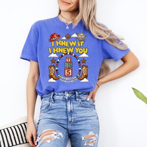May include: A blue short-sleeve t-shirt featuring a graphic print with the text I KNEW IT I KNEW YOU, a red cowboy hat, a sheriff badge, two brown cowboy boots, and a central stack of three alphabet blocks with the letters T, S, and the number 5.