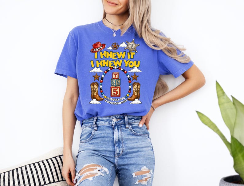 May include: A blue short-sleeve t-shirt featuring a graphic print with the text I KNEW IT I KNEW YOU, a red cowboy hat, a sheriff badge, two brown cowboy boots, and a central stack of three alphabet blocks with the letters T, S, and the number 5.