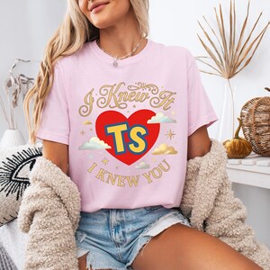 May include: A light pink short-sleeve t-shirt featuring a graphic design with a large red heart, the yellow and blue letters TS, and the text I KNEW IT I KNEW YOU in gold cursive lettering, accented with small clouds and star shapes.