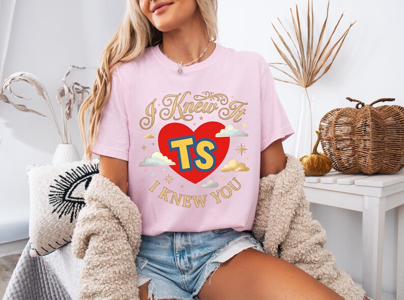 May include: A light pink short-sleeve t-shirt featuring a graphic design with a large red heart, the yellow and blue letters TS, and the text I KNEW IT I KNEW YOU in gold cursive lettering, accented with small clouds and star shapes.