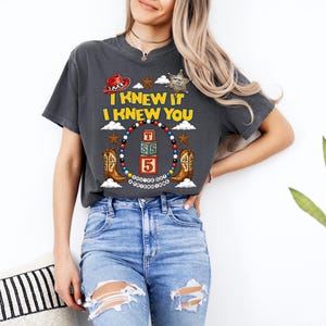 May include: A charcoal gray short-sleeve t-shirt featuring a graphic print with the text I KNEW IT I KNEW YOU, a red cowboy hat, a sheriff star, cowboy boots, and a circular frame of colorful beads surrounding stacked alphabet blocks.