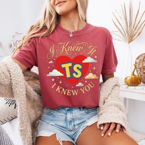 May include: A red short-sleeved t-shirt featuring a graphic of a large red heart with the yellow letters TS inside. The text I KNEW IT is printed in gold above the heart, and I KNEW YOU is printed in gold below, surrounded by clouds and stars.