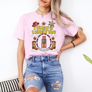 May include: A light pink short-sleeve t-shirt featuring a graphic print with the text "I KNEW IT I KNEW YOU" above a circular beaded border, cowboy boots, a red hat, a sheriff badge, and stacked blocks with the letters "TSS" and the number "5".
