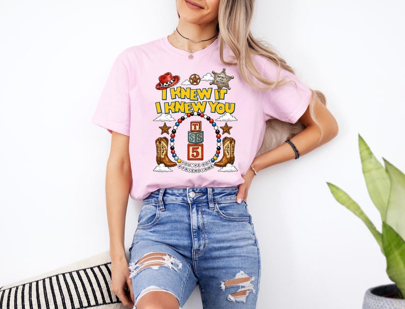 May include: A light pink short-sleeve t-shirt featuring a graphic print with the text "I KNEW IT I KNEW YOU" above a circular beaded border, cowboy boots, a red hat, a sheriff badge, and stacked blocks with the letters "TSS" and the number "5".