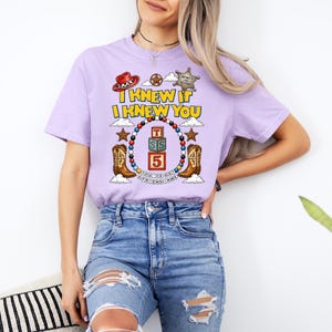 May include: A light purple short-sleeve t-shirt featuring a graphic print with the text I KNEW IT I KNEW YOU, a sheriff badge, a cowboy hat, two cowboy boots, and a central stack of three blocks labeled T, S, and 5 surrounded by colorful beads.