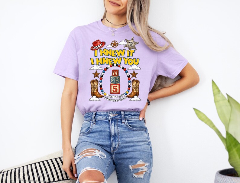 May include: A light purple short-sleeve t-shirt featuring a graphic print with the text I KNEW IT I KNEW YOU, a sheriff badge, a cowboy hat, two cowboy boots, and a central stack of three blocks labeled T, S, and 5 surrounded by colorful beads.
