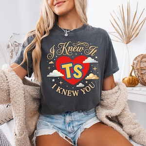 May include: A charcoal grey short-sleeve t-shirt featuring a graphic design with a red heart, the yellow letters TS, and the text I KNEW IT I KNEW YOU in gold script. The design includes small white clouds and gold stars surrounding the central heart.
