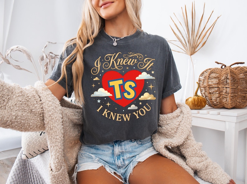 May include: A charcoal grey short-sleeve t-shirt featuring a graphic design with a red heart, the yellow letters TS, and the text I KNEW IT I KNEW YOU in gold script. The design includes small white clouds and gold stars surrounding the central heart.