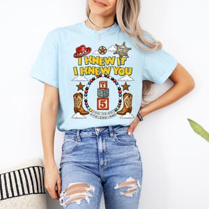 May include: A light blue t-shirt featuring a graphic print with the text I KNEW IT I KNEW YOU, a sheriff badge, a red cowboy hat, two brown cowboy boots, and a central stack of three alphabet blocks labeled T, S, and 5, surrounded by beads.