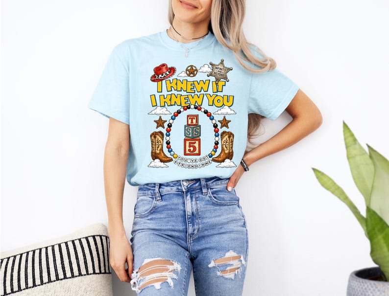 May include: A light blue t-shirt featuring a graphic print with the text I KNEW IT I KNEW YOU, a sheriff badge, a red cowboy hat, two brown cowboy boots, and a central stack of three alphabet blocks labeled T, S, and 5, surrounded by beads.