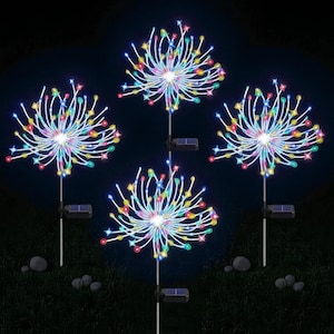 Può includere: Quattro luci da giardino a energia solare a forma di fuochi d'artificio. Ogni luce ha una luce bianca centrale con fili LED colorati e radianti. Le luci sono su pali di metallo con piccoli pannelli solari, su sfondo scuro.