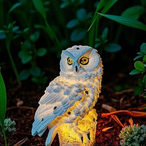 May include: A detailed, illuminated owl statue with a light blue and white color scheme, perched on a tree stump. The owl has large yellow eyes and is surrounded by green foliage, creating a natural garden ornament.