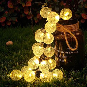 Könnte beinhalten: Eine Lichterkette mit kugelförmigen LED-Leuchten mit strukturiertem, blasenartigem Aussehen, die ein warmes gelbes Leuchten ausstrahlen. Die Lichter sind auf grünem Gras angeordnet. Ein dunkles Glasgefäß mit einer Schnur ist im Hintergrund.