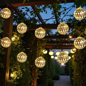 Puede incluir: Cadena de luces decorativas con globos dorados. Cada globo presenta un patrón calado detallado y emite una luz cálida. La cadena de luces está suspendida de un cable oscuro, en un entorno exterior con vegetación y una pérgola de madera.