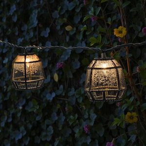 Könnte beinhalten: Lichterkette mit zwei dekorativen, käfigartigen Laternen. Die schwarzen Laternen haben eine strukturierte Oberfläche, mit warmem, gelbem Licht, das von innen kommt. Der Hintergrund zeigt üppiges grünes Laub mit kleinen Blüten.