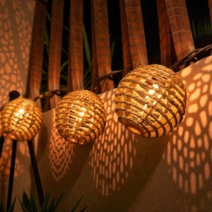 May include: A string of spherical rattan lights, each emitting a warm light. The lights are suspended against a light-coloured wall and wooden fence, casting patterned shadows. The overall effect is a warm and inviting ambiance, perfect for outdoor settings.