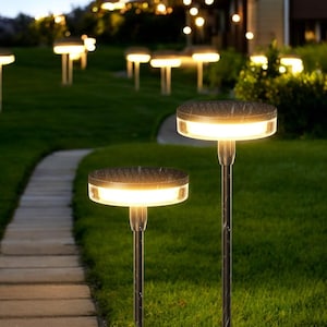 Pode incluir: Luzes solares para jardim com um design moderno. As luzes têm um topo escuro e texturizado e uma fonte de luz circular e brilhante. Estão colocadas num relvado verde com um caminho de pedra, criando um ambiente acolhedor para um jardim. As luzes têm cerca de 60 cm de altura.