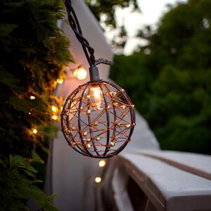 Könnte beinhalten: Dekorative Außen-Lichterkette mit warmem Licht. Die Leuchte hat ein kugelförmiges Metallkäfig-Design mit kleinen, goldenen Akzenten. Das Licht wird an einem dunklen Draht aufgehängt und schafft eine gemütliche Atmosphäre für den Außenbereich.