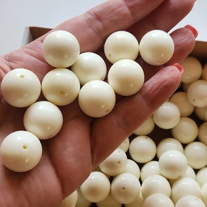 May include: A box filled with white round beads. The beads are smooth and have a slight shine. The beads are in a hand, and the box is open.