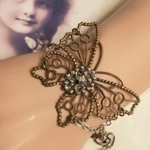 May include: A delicate bronze butterfly bracelet with a crystal center and a dangling charm. The bracelet is on a mannequin's wrist.