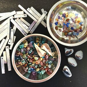 May include: A silver bowl filled with colorful gemstones, a silver bowl filled with colorful seashells, and a pile of silver metal bars.