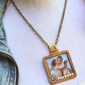 May include: A vintage-style gold-tone necklace featuring a rectangular pendant with a framed cherub painting. The pendant is adorned with a decorative crystal flower and a delicate chain.
