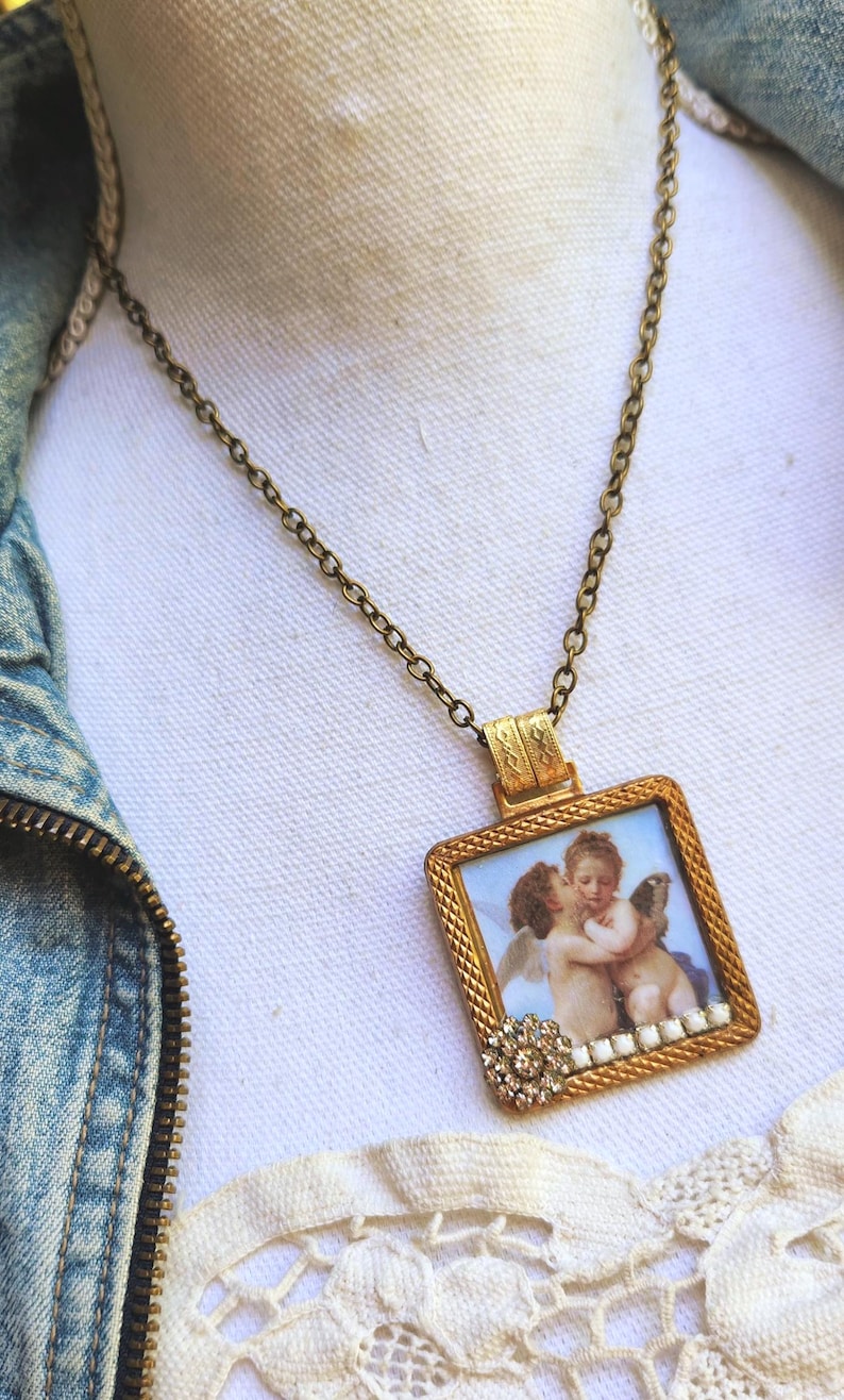 May include: A vintage-style gold-tone necklace featuring a rectangular pendant with a framed cherub painting. The pendant is adorned with a decorative crystal flower and a delicate chain.