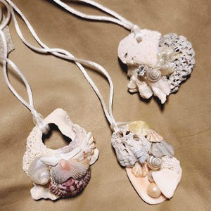 May include: Three seashell necklaces with white cords. Each necklace features a unique arrangement of natural seashells in various shapes, sizes, and colors, including white, beige, and hints of pink and purple. Some necklaces have decorative elements.