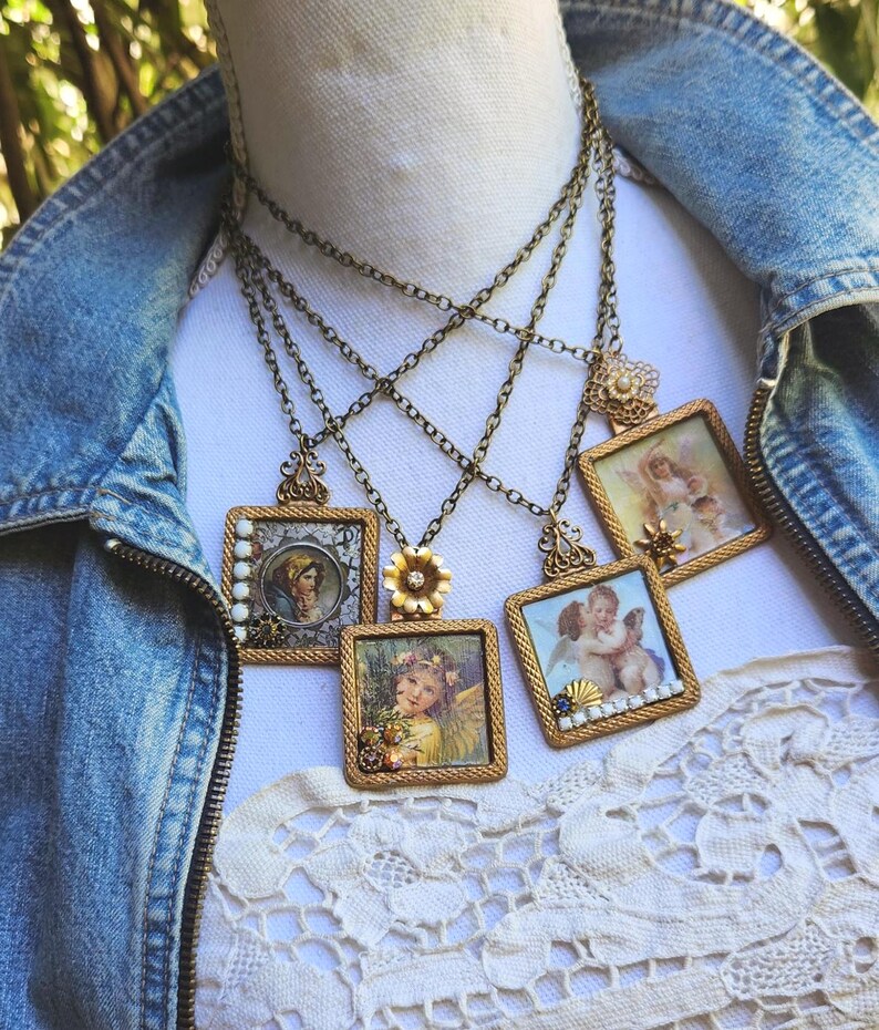 May include: A collection of vintage-style necklaces with square pendants featuring angel and portrait illustrations. The pendants are framed in ornate gold-toned metal and hang from delicate chains. Some pendants have floral or bead accents.