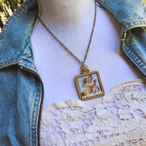 May include: A vintage gold-toned necklace with a square pendant featuring a painting of two cherubs. The pendant is framed with a gold border and small white and blue accents. The necklace is displayed on a white surface.