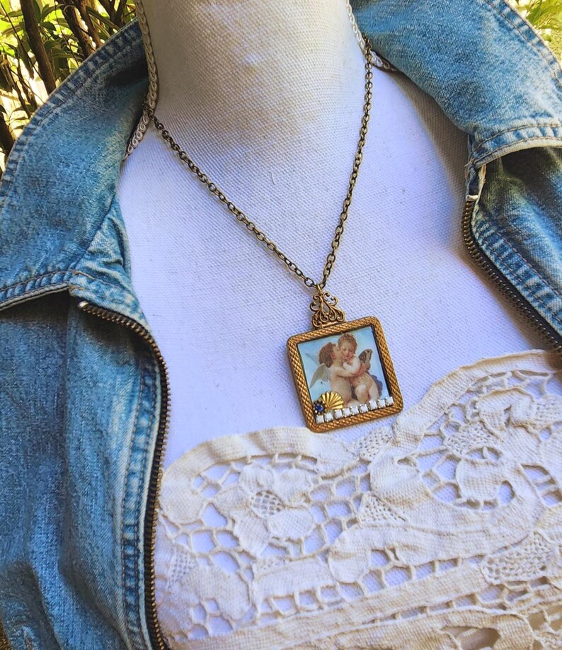 May include: A vintage gold-toned necklace with a square pendant featuring a painting of two cherubs. The pendant is framed with a gold border and small white and blue accents. The necklace is displayed on a white surface.