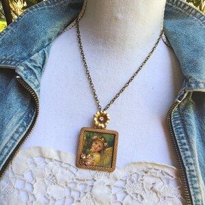 May include: A vintage-style necklace featuring a square pendant with a picture of an angel. The pendant is framed in gold-tone metal and adorned with small crystals. The necklace has a delicate chain and a flower-shaped clasp.