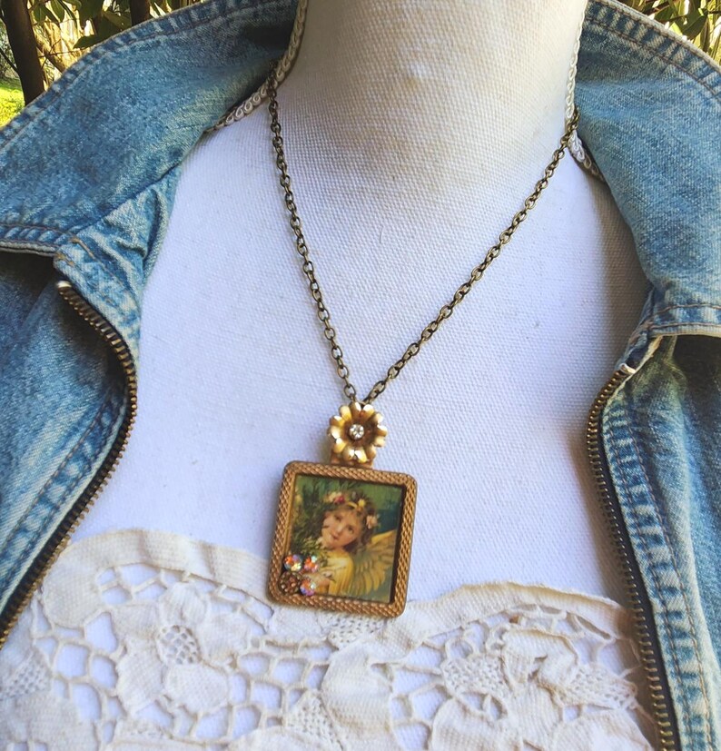 May include: A vintage-style necklace featuring a square pendant with a picture of an angel. The pendant is framed in gold-tone metal and adorned with small crystals. The necklace has a delicate chain and a flower-shaped clasp.