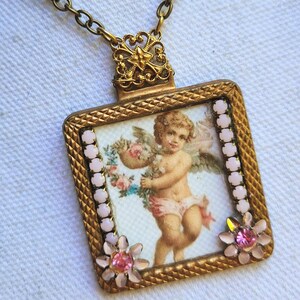 May include: A vintage-style gold-toned pendant necklace. The square pendant features a cherub illustration holding flowers, framed with pink accents and floral embellishments. The necklace has a delicate chain.