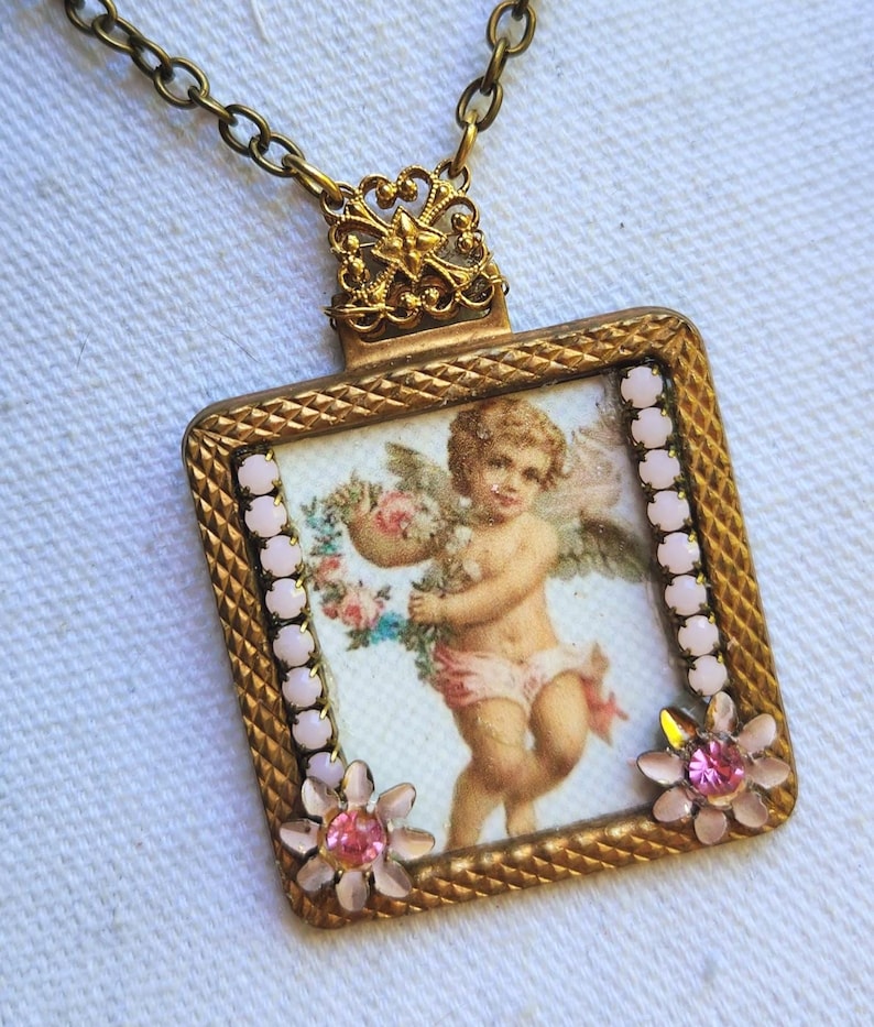 May include: A vintage-style gold-toned pendant necklace. The square pendant features a cherub illustration holding flowers, framed with pink accents and floral embellishments. The necklace has a delicate chain.