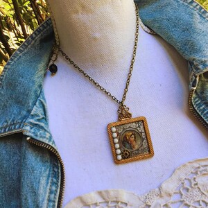 May include: A vintage-style pendant necklace with a square, gold-toned frame. The pendant features a portrait of a person in a headscarf, surrounded by white beads. The necklace is worn over a denim jacket.