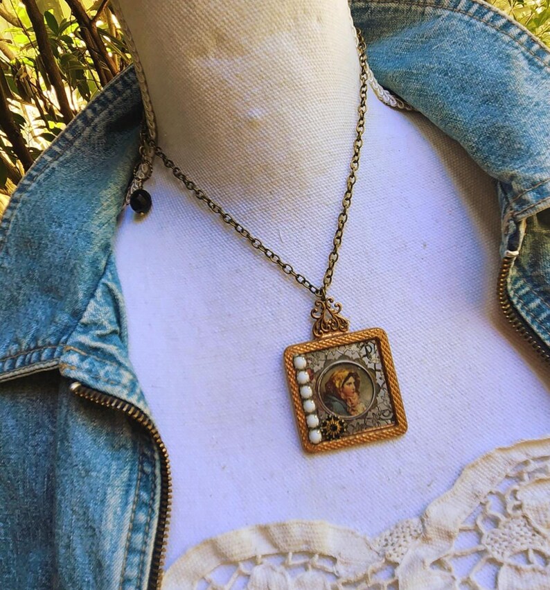 May include: A vintage-style pendant necklace with a square, gold-toned frame. The pendant features a portrait of a person in a headscarf, surrounded by white beads. The necklace is worn over a denim jacket.