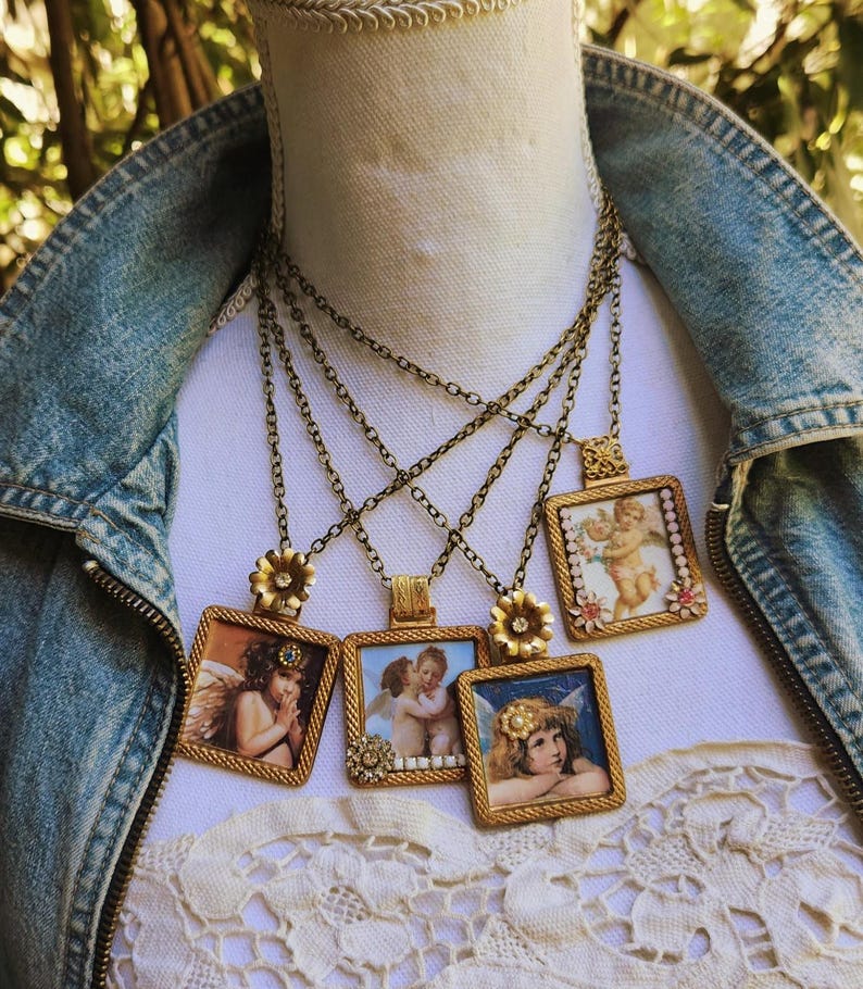 May include: A collection of antique-style necklaces featuring square pendants with cherub artwork. The pendants are framed in gold-colored metal and hang from delicate chains. Each necklace has a unique cherub image.