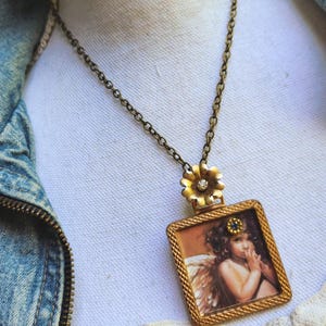 May include: A vintage-style necklace with a rectangular pendant featuring an angel illustration. The pendant is framed in gold and hangs from a bronze chain. A gold flower with a central gem connects the pendant to the chain.