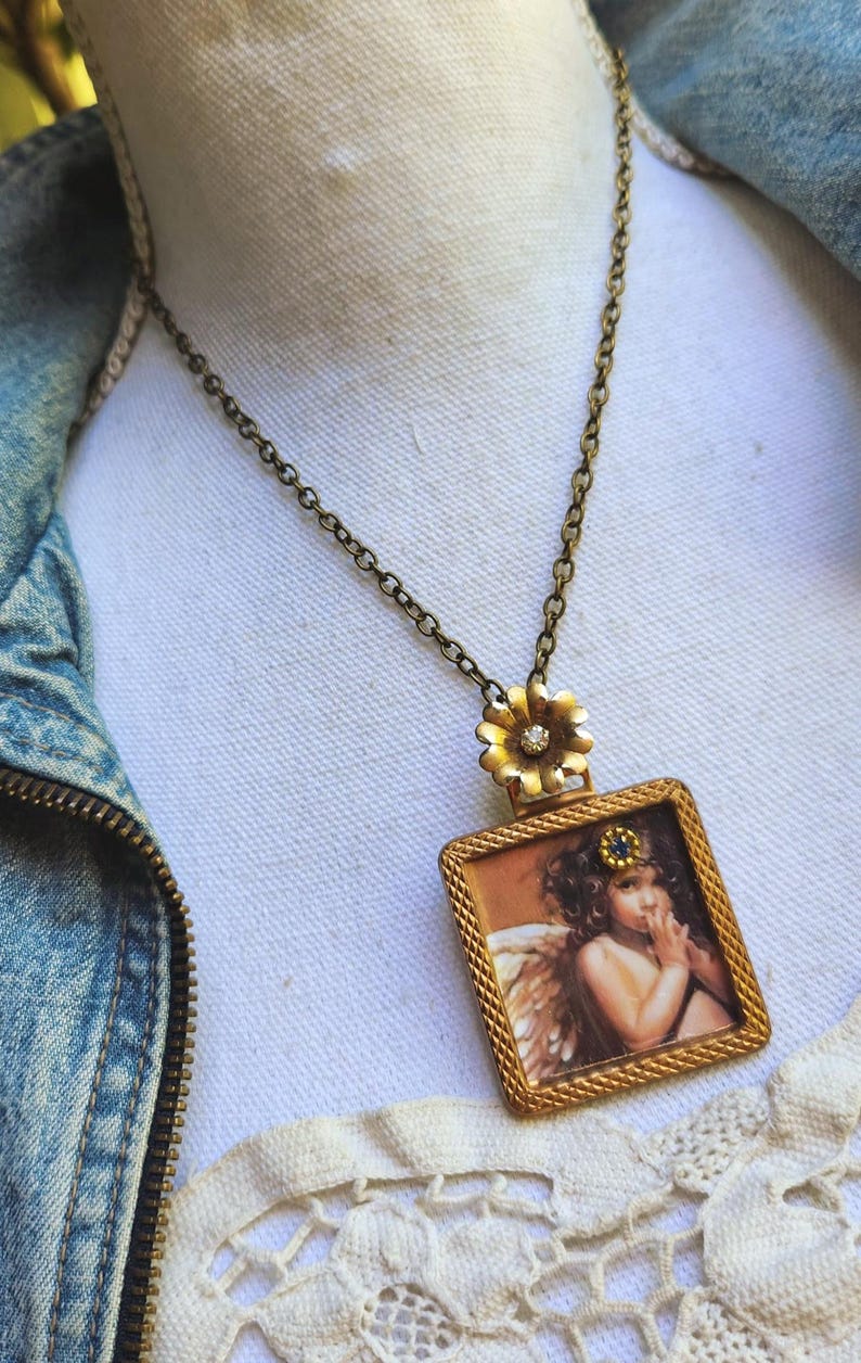 May include: A vintage-style necklace with a rectangular pendant featuring an angel illustration. The pendant is framed in gold and hangs from a bronze chain. A gold flower with a central gem connects the pendant to the chain.