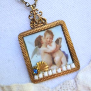 May include: A vintage-style pendant necklace with a square, gold-toned frame. The frame holds a miniature painting of cherubs. The pendant is adorned with a blue gemstone, a gold seashell, and white beads. The chain is a matching gold color.
