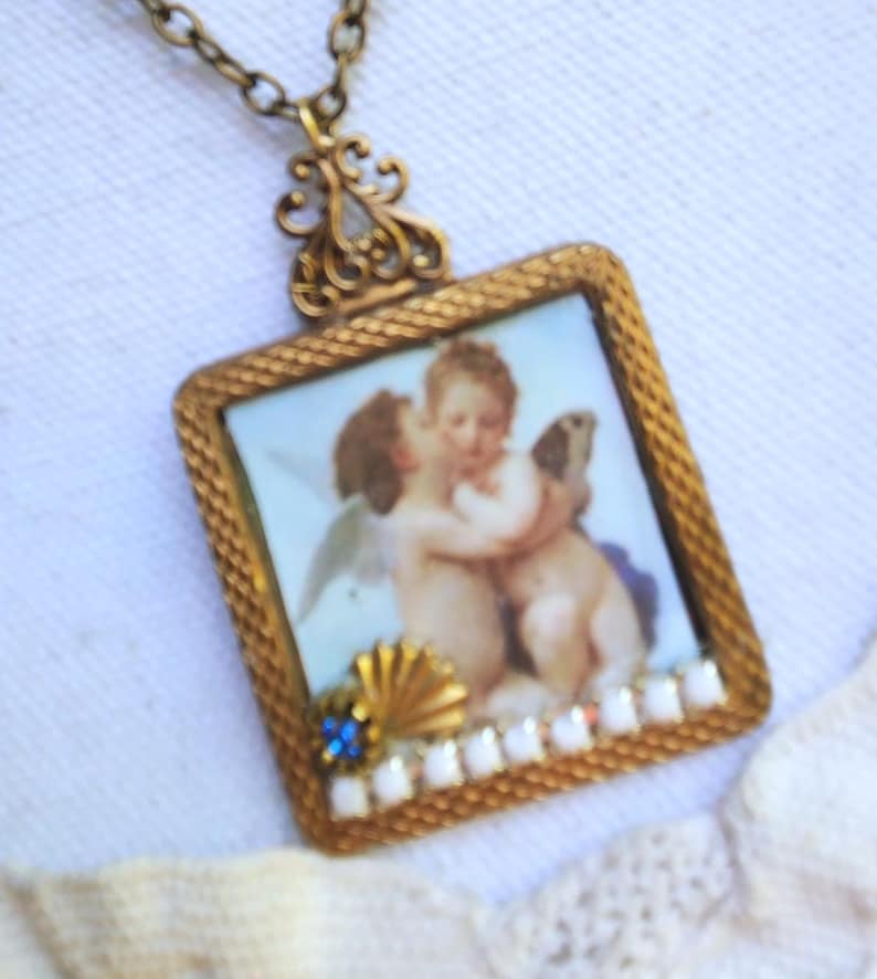 May include: A vintage-style pendant necklace with a square, gold-toned frame. The frame holds a miniature painting of cherubs. The pendant is adorned with a blue gemstone, a gold seashell, and white beads. The chain is a matching gold color.